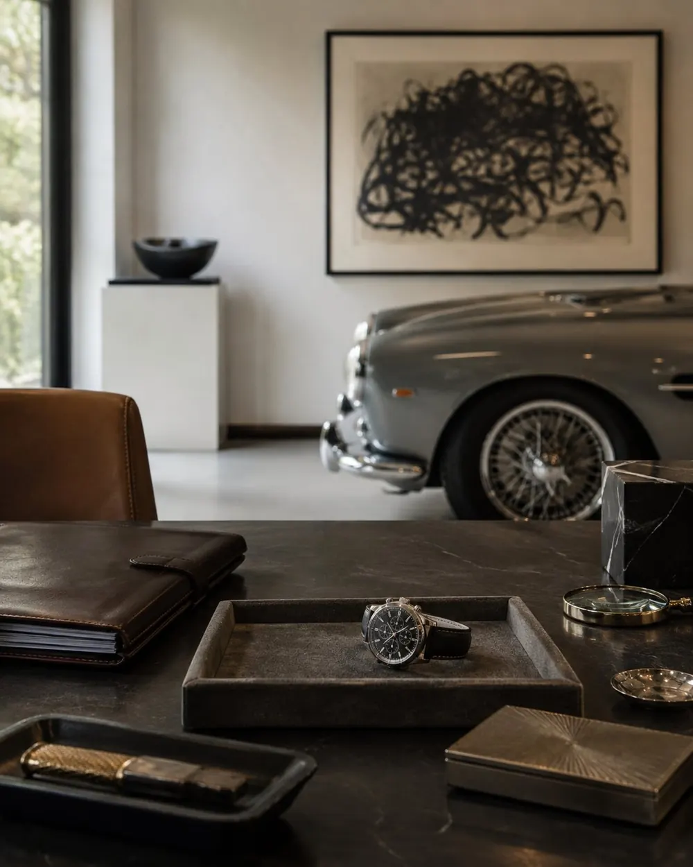 A desk with a watch and leather trays, a classic car in the room beyond